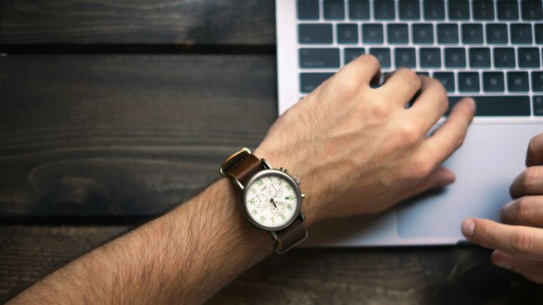 Forearm with a wristwatch resting on a laptop on a dark wooden desk